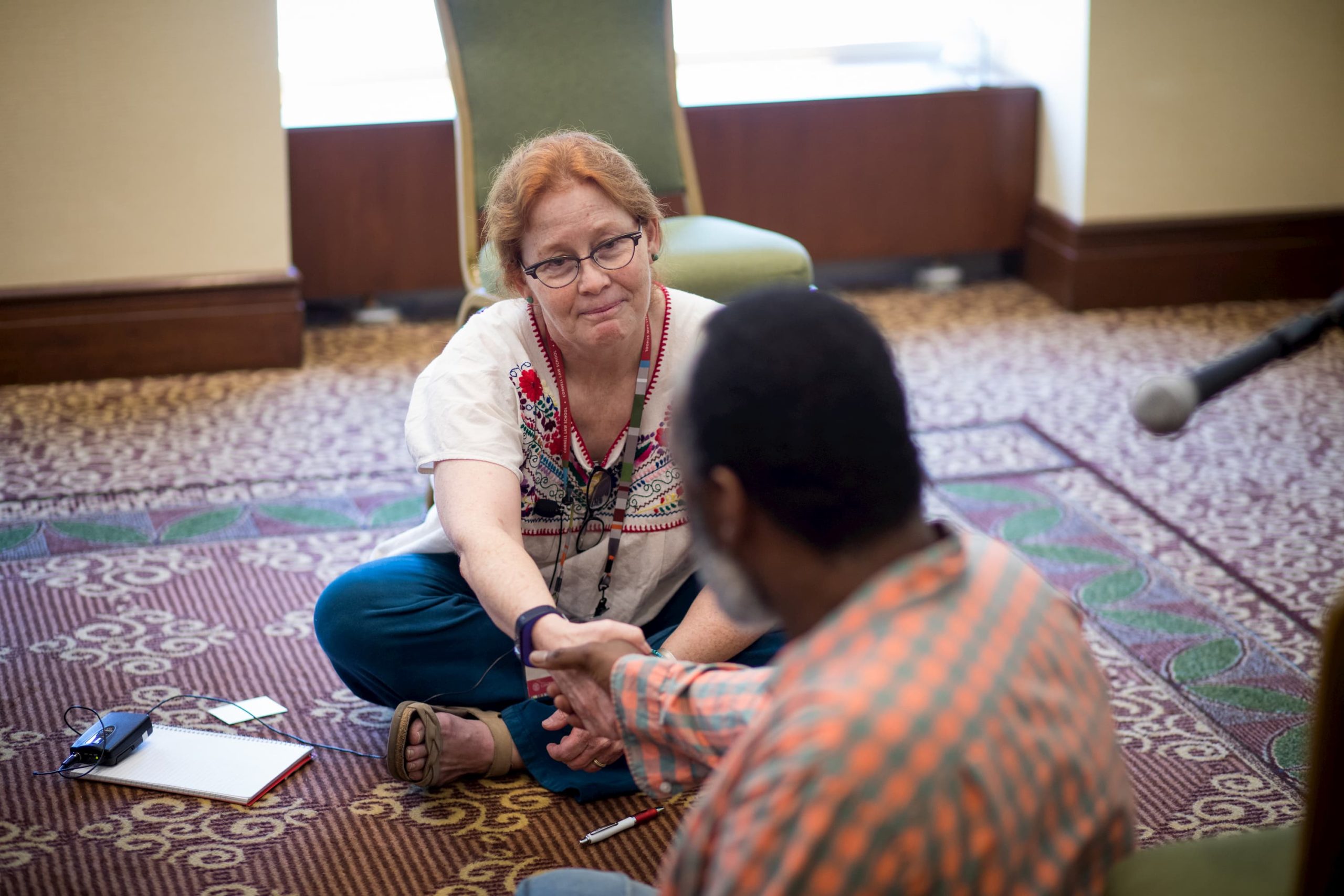 A woman with glasses and in a white shirt with embroidery is sitting on the ground, holding hands with a man in an orange plaid shirt.