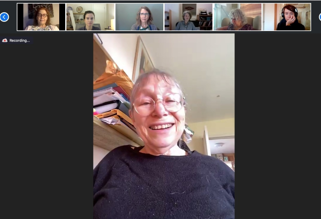 A woman wearing glasses and a gray shirt speaks during a video conference. Six people, their pictures smaller, listen.