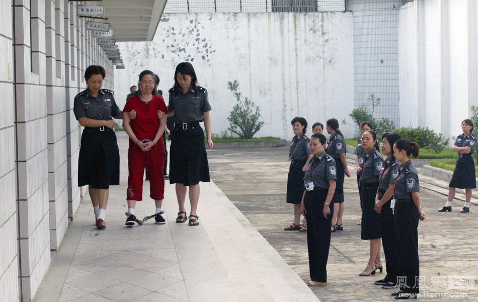 Two guards walk beside a woman who is wearing red and whose ankles and wrists are chained. A group of guards stands next to them and watches as they walk.